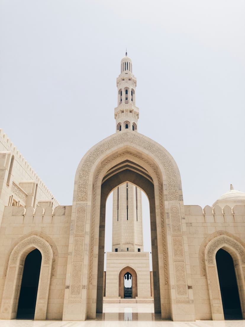 Tall stone minaret rises between ornate pointed arches under a bright blue sky at the Omani Grand Mosque.