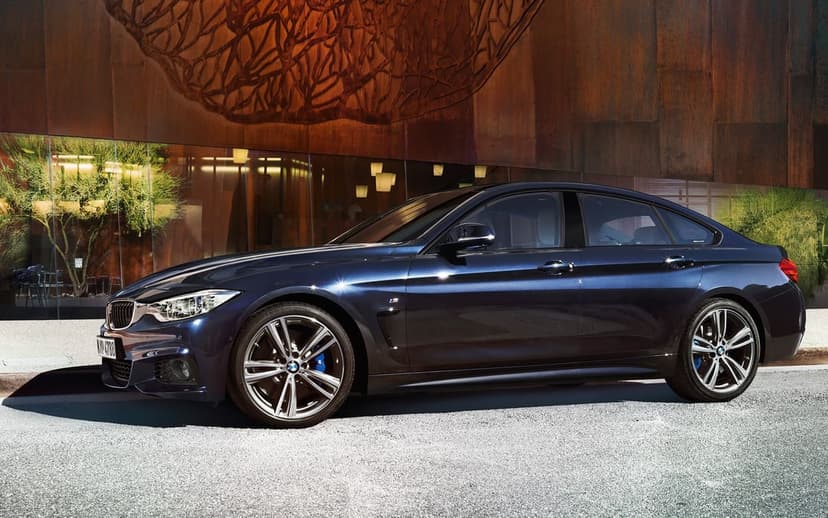 Dark blue BMW 4 Series Gran Coupe parked in front of a textured metal wall and glass windows on a sunny day.