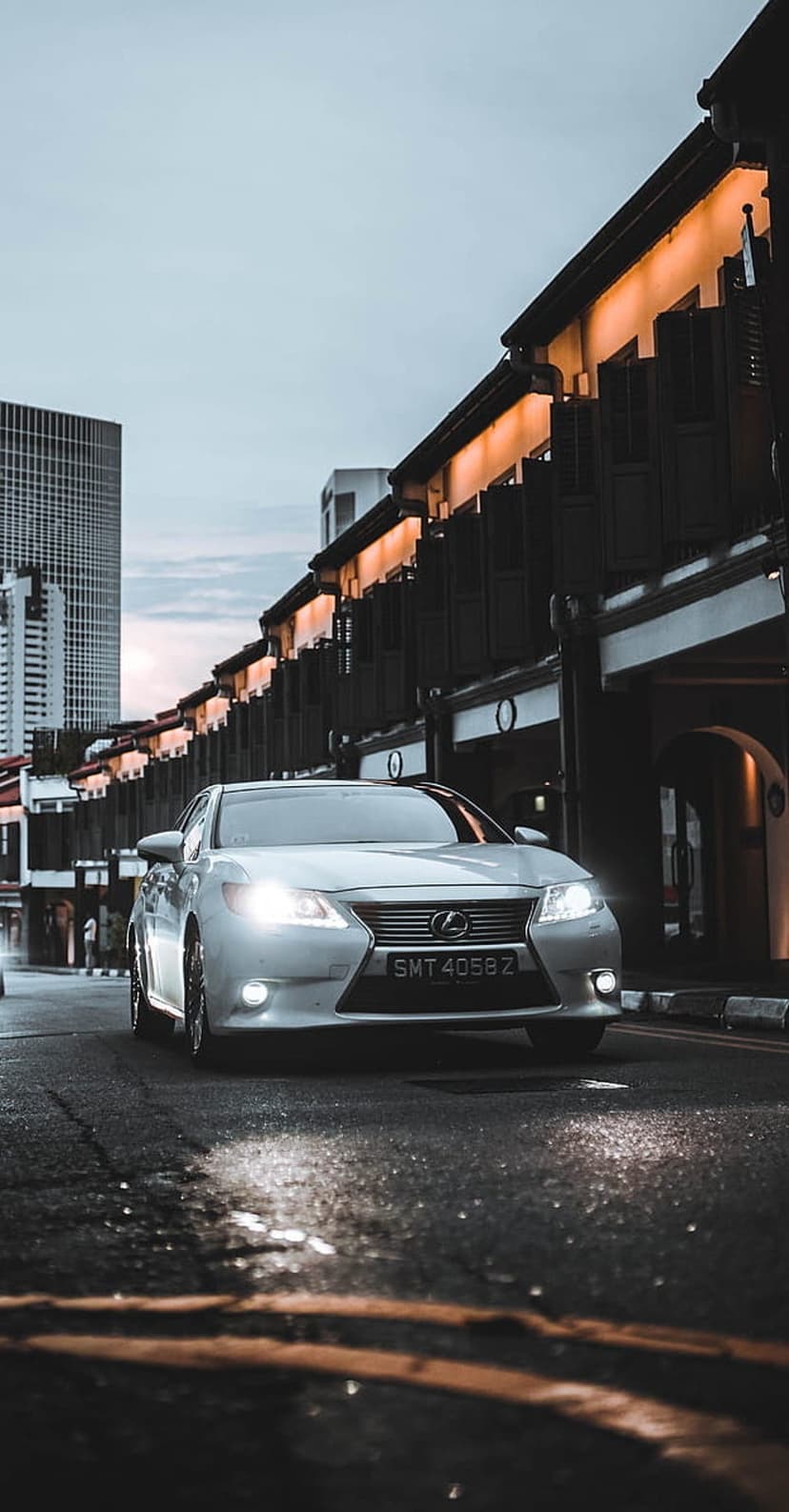 Silver Lexus sedan parked on a wet street with glowing headlights, urban architecture, and a skyscraper at dusk.