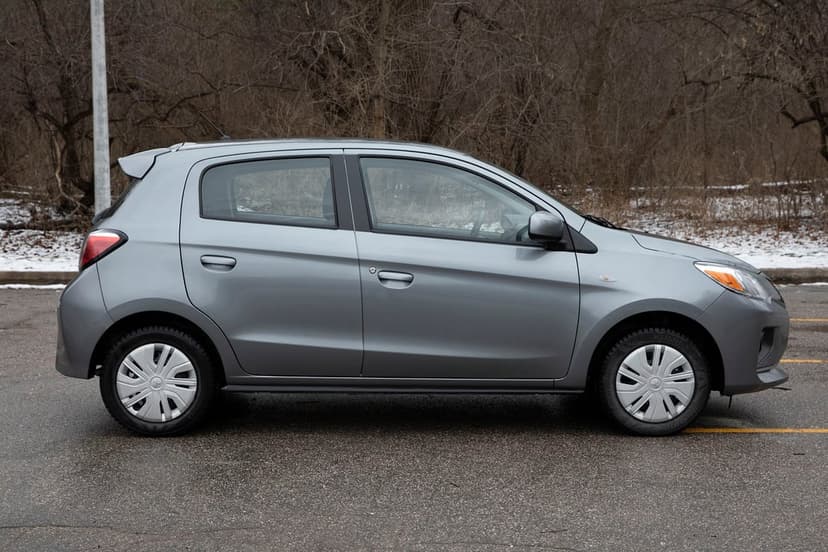 Silver Mitsubishi Mirage hatchback side profile parked on wet asphalt against a wintry forest background.