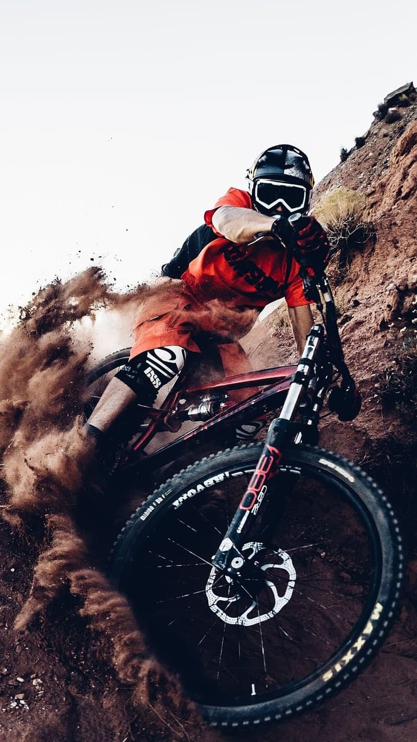 Mountain biker in a red jersey navigates a steep rocky trail while kicking up a thick cloud of dust and debris.