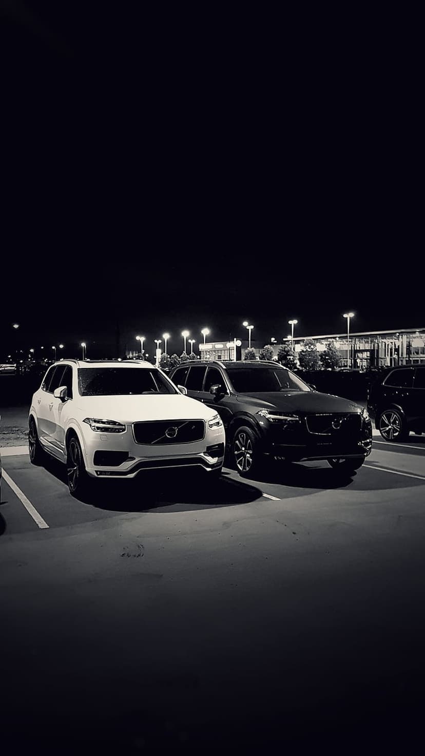 Volvo XC90 SUVs parked in a dark lot at night with glowing headlights and a lit modern building in the background.