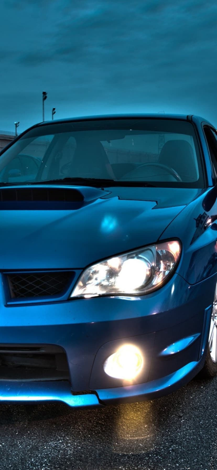 Blue Subaru Impreza front headlight and hood scoop glowing brightly in a low-light evening setting.