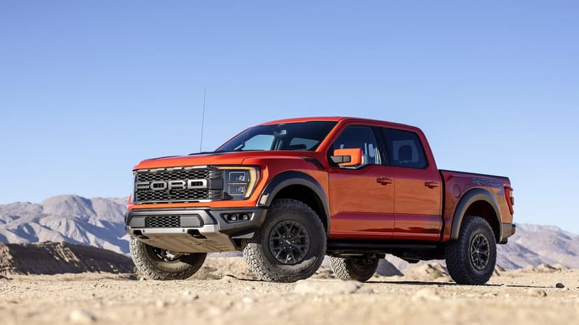 Orange Ford F-150 Raptor truck on sandy desert ground with mountains and a clear blue sky in the background.
