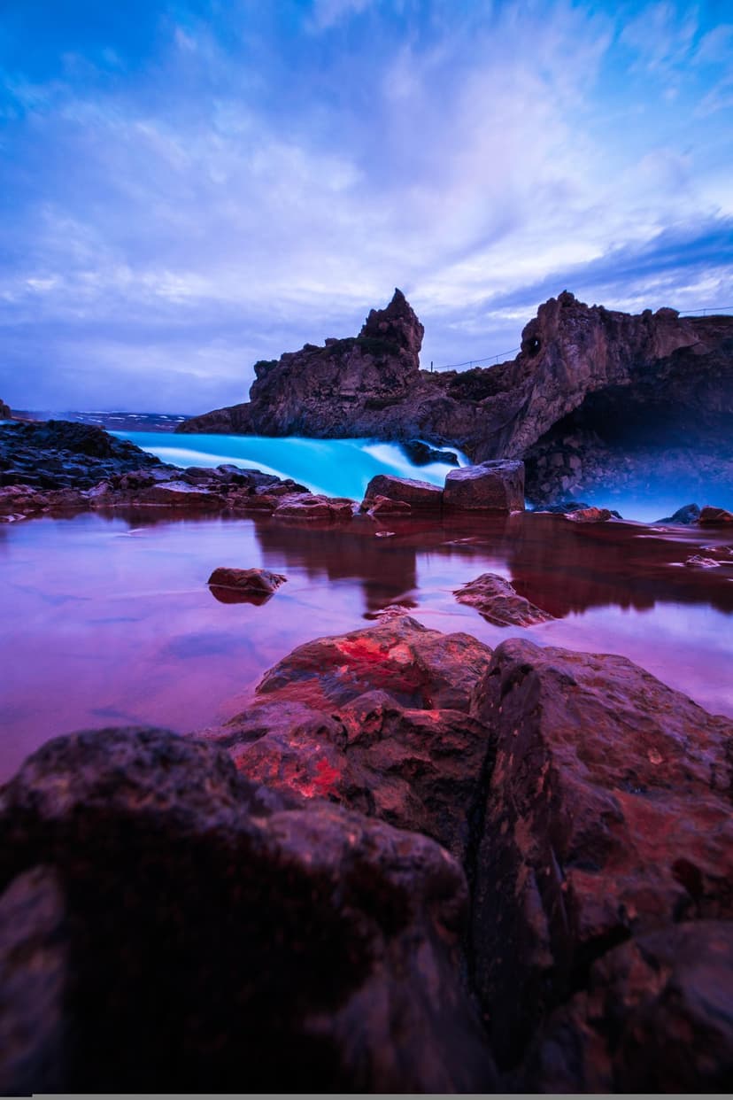 Turquoise waterfall cascading into a purple reflection pool surrounded by dark jagged rocks under a twilight sky.
