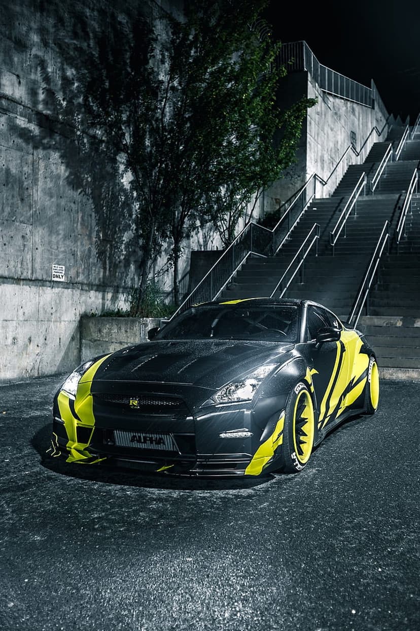Black Nissan GT-R with yellow graphics and wheels parked on wet asphalt next to an illuminated urban staircase.