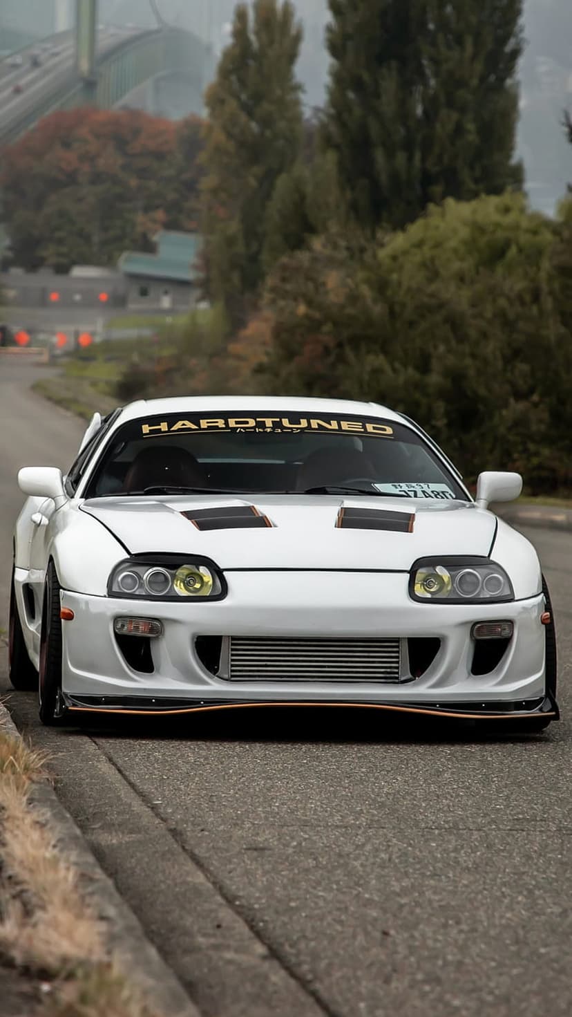White Toyota Supra with a slammed stance and Hardtuned banner parked on a misty street near a blurred bridge.