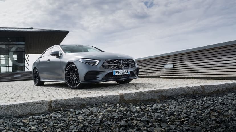 Matte grey Mercedes CLS sedan parked on a cobblestone driveway in front of a modern building with a cloudy sky.