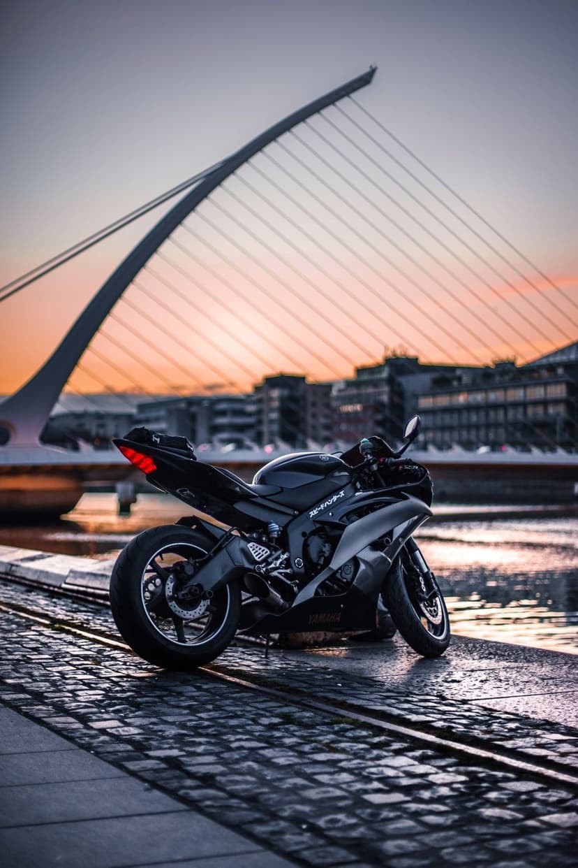 Black sportbike on cobblestones with the harp-shaped Samuel Beckett Bridge under a glowing sunset in Dublin.