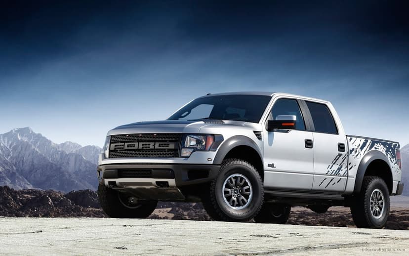 Silver Ford F-150 SVT Raptor pickup truck in a desert with snowy mountains under a deep blue sky.