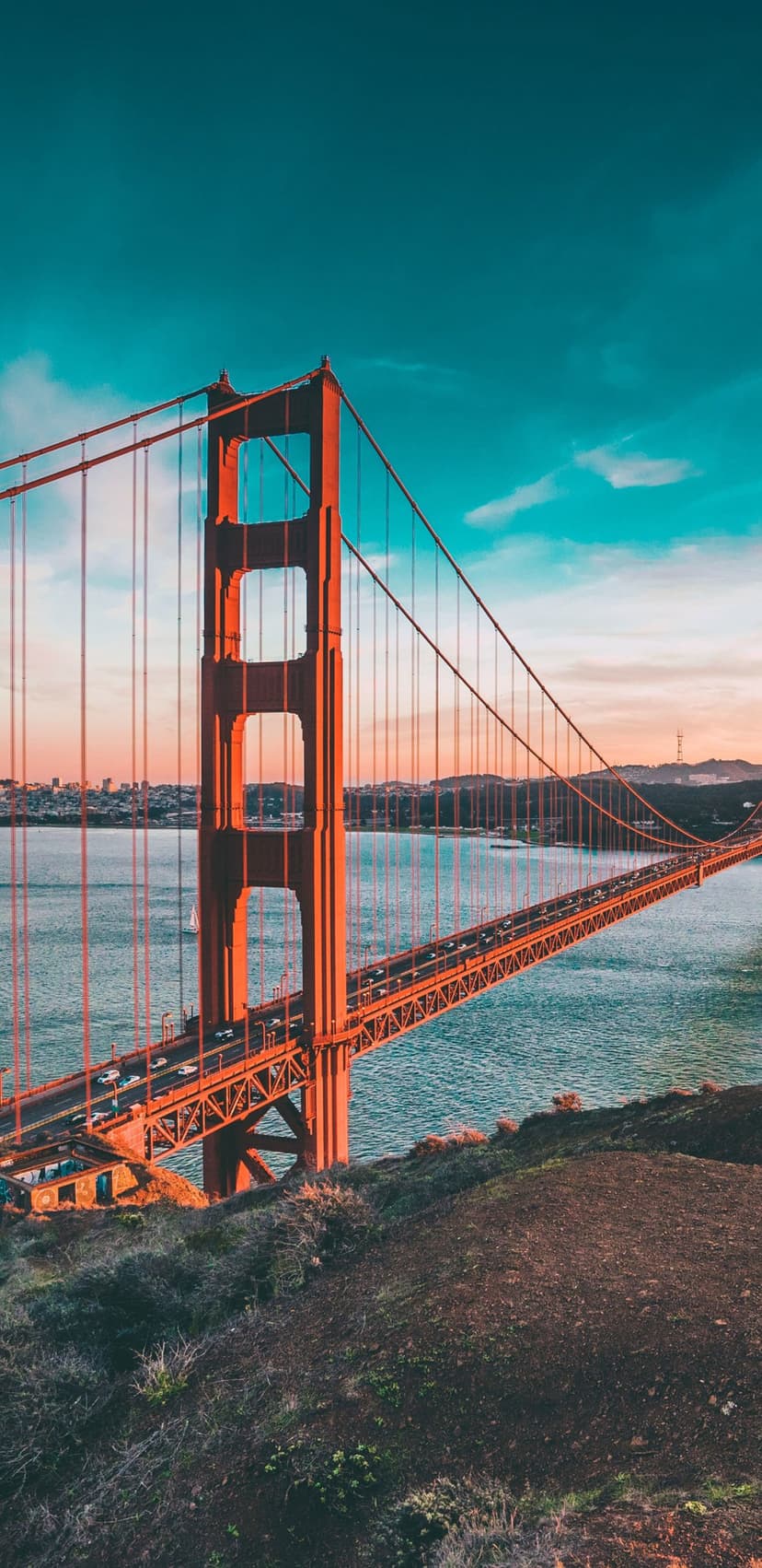 Golden Gate Bridge towers and cables rise above blue bay waters under a teal and orange sunset sky at dusk.