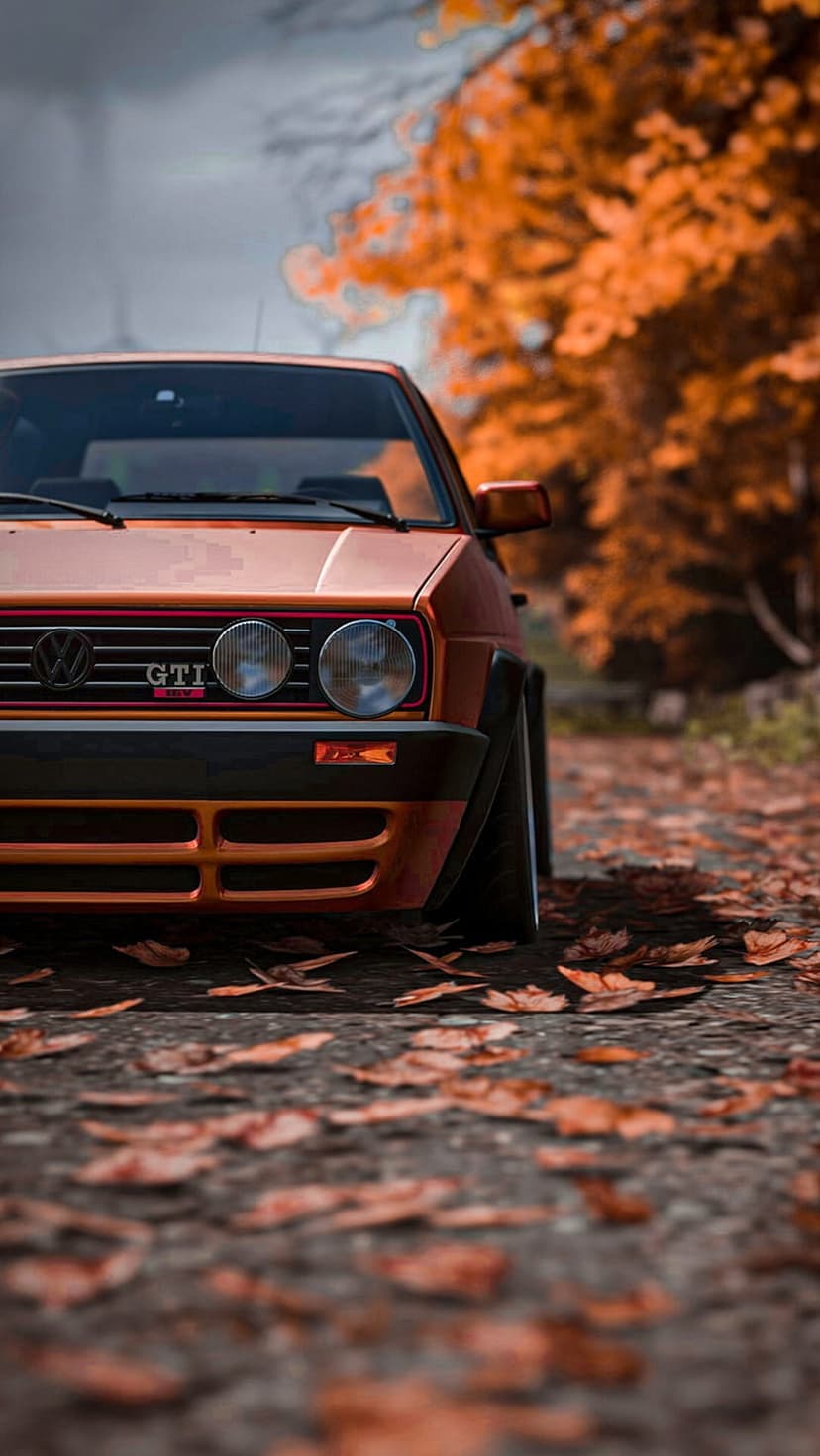 Orange Volkswagen Golf GTI Mk2 parked on a road covered in autumn leaves with a blurred forest background.