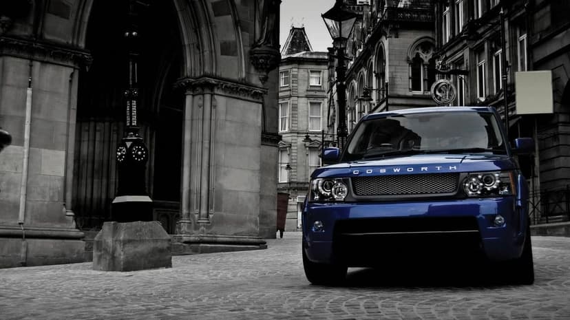 Blue Range Rover Sport with Cosworth lettering on a grayscale cobblestone street by historic stone buildings.