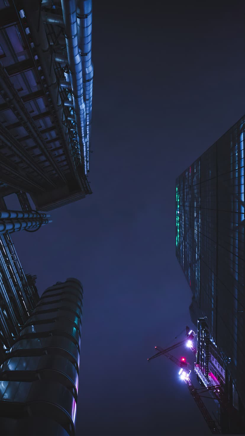 Tall skyscrapers with glowing blue windows tower over a construction crane against a dark blue midnight sky.