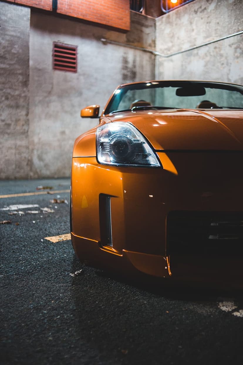 Orange Nissan 350Z headlight and fender reflect ambient night light against an urban concrete wall background.