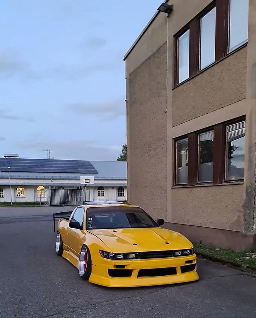 Yellow Nissan Silvia S13 with a large rear spoiler and body kit parked on asphalt near a building with solar panels.