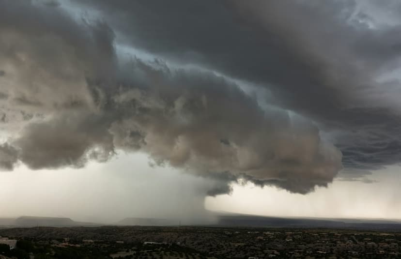 Dark shelf clouds hover above a hazy city horizon with heavy rain falling from the thick gray storm base.