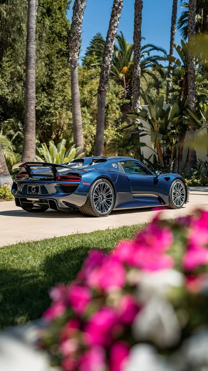 Blue Porsche 918 Spyder parked on a driveway lined with palm trees and pink flowers under a clear blue sky.