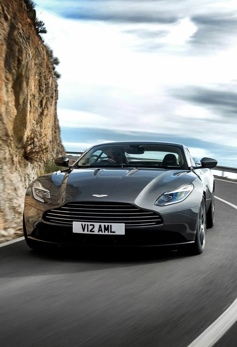 Dark grey Aston Martin convertible driving on a winding mountain road next to a steep cliff under a cloudy sky.