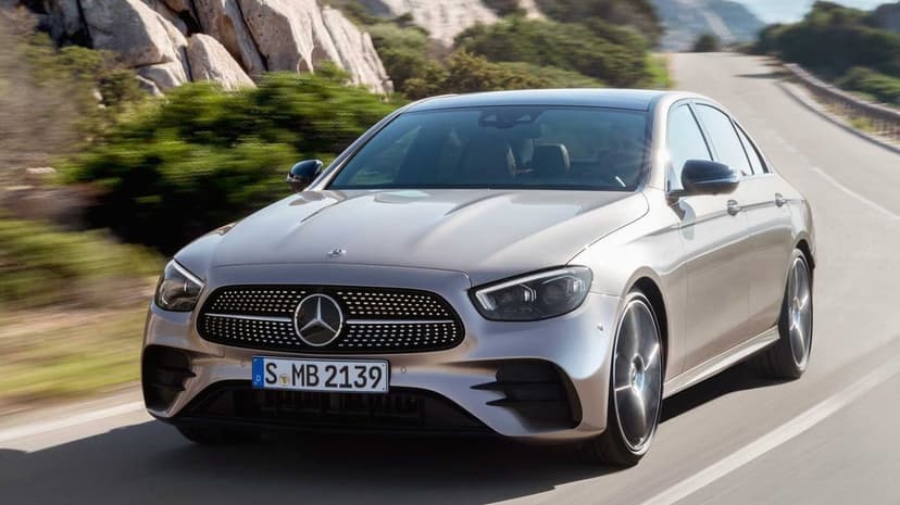Silver Mercedes-Benz sedan driving on a winding coastal road with motion blur and rocky cliffs background.