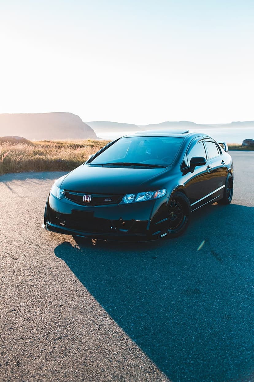 Black Honda Civic SI parked on an asphalt coastal road during sunset with rolling hills and water in the background.