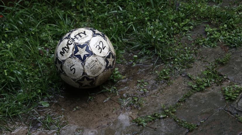 Weathered soccer ball with black stars sits on wet mossy paving stones and grass during an overcast day.