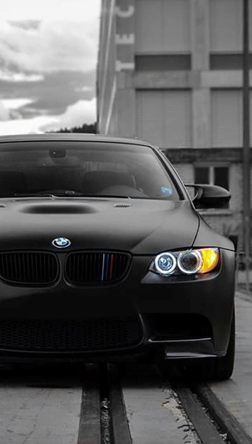 Matte black BMW M3 with yellow angel eye headlights sits on metal tracks against a desaturated urban skyline.