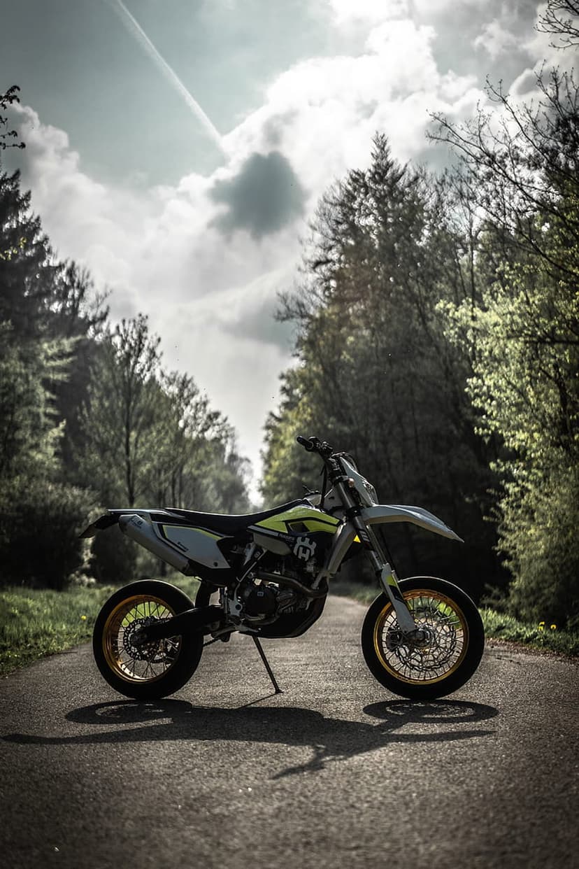 White and yellow Husqvarna motorcycle on an asphalt road in a green forest under dramatic clouds and sunbeams.