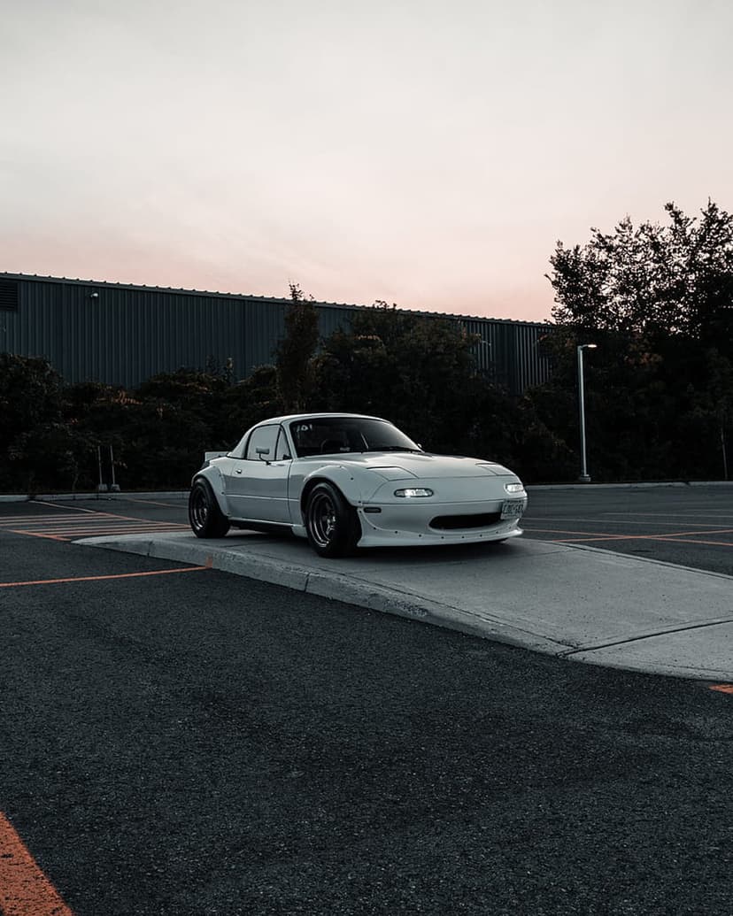White widebody Mazda Miata parked on a concrete slope during a golden sunset with dark industrial buildings.