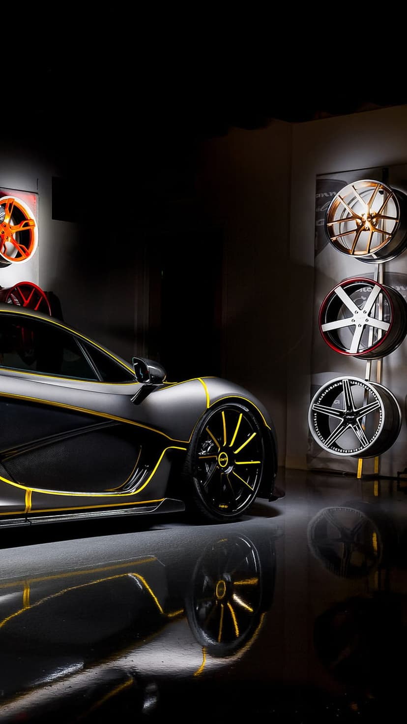 McLaren P1 supercar in matte black with yellow racing stripes parked in a studio with custom wheels on racks.