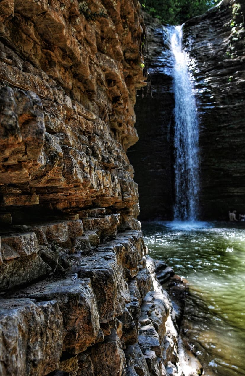 Slender waterfall cascades into a clear green pool surrounded by layered rock walls in a sunlit mountain gorge.