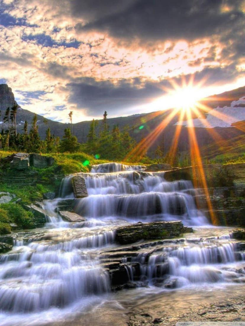 Multi-tiered waterfall flows over mossy rocks under a cloudy sky with bright sun rays during golden hour.