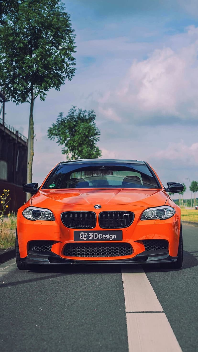 Orange BMW M5 with a 3DDesign body kit parked on a road under a cloudy sky with a bridge in the distance.
