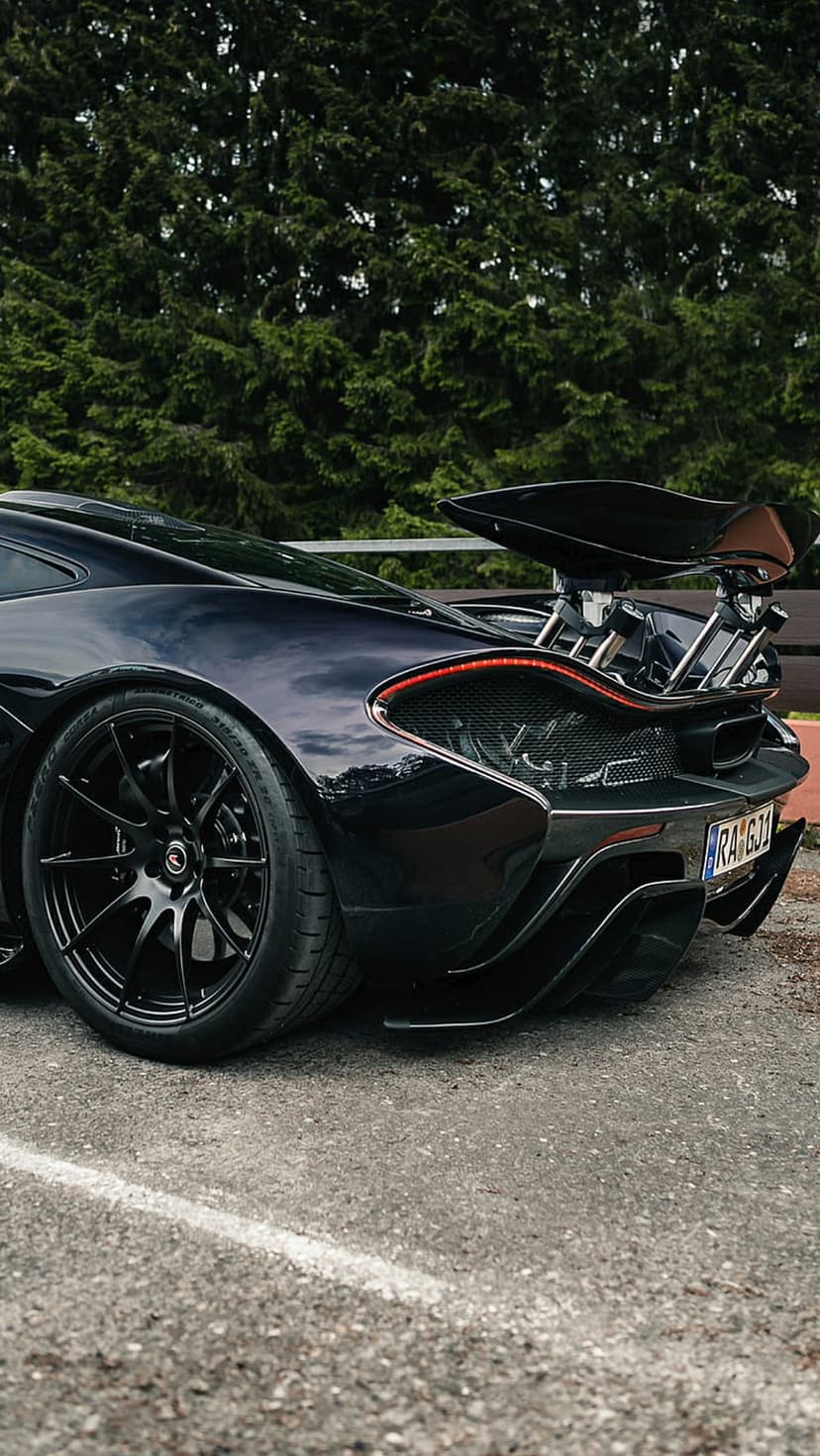 McLaren P1 rear view with a large spoiler and LED lights parked on a track against a backdrop of green trees.