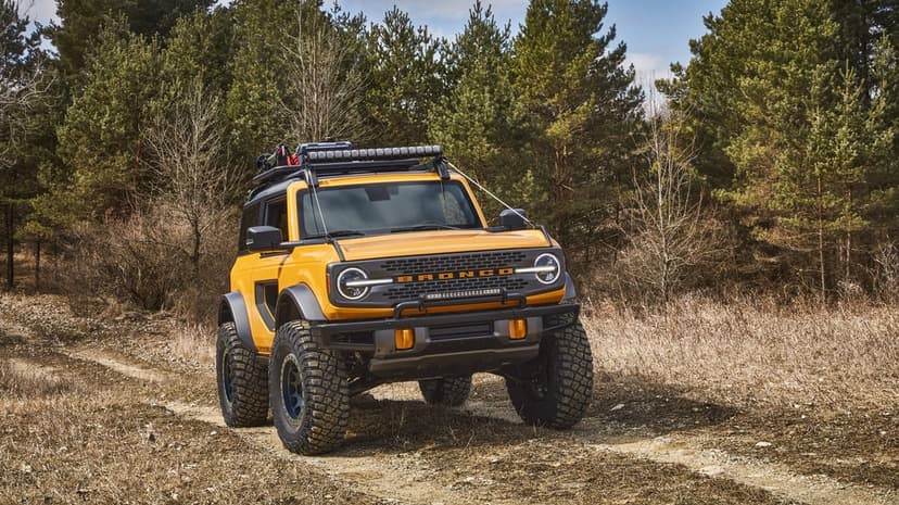 Yellow Ford Bronco with off-road tires and roof rack on a dirt trail in a dense forest during daylight.