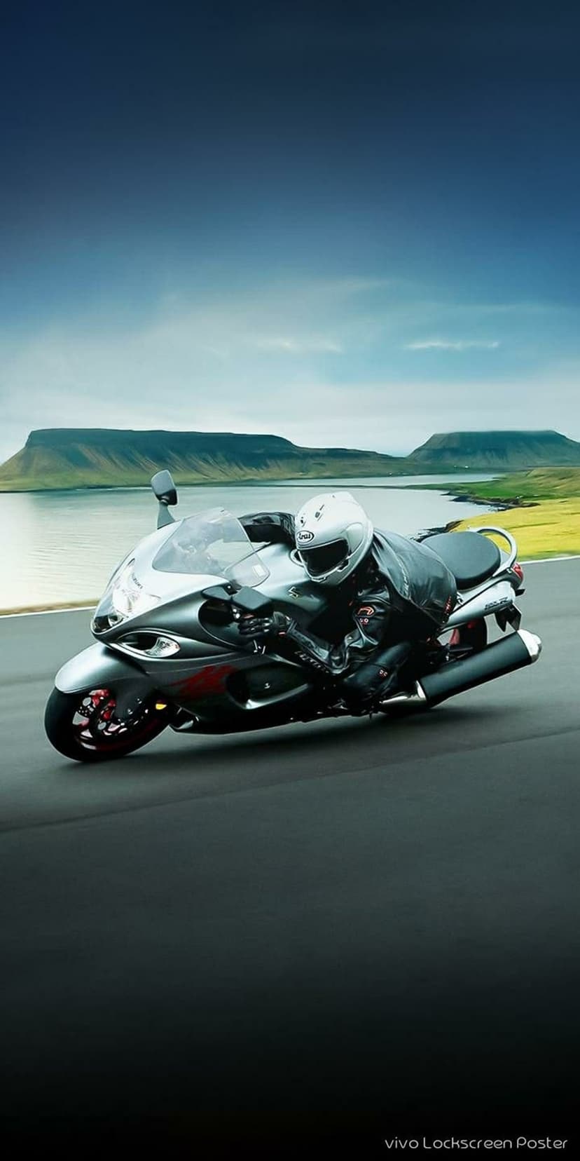 Motorcyclist in silver gear leans a sportbike into a mountain road turn beside a calm blue lake under clouds.
