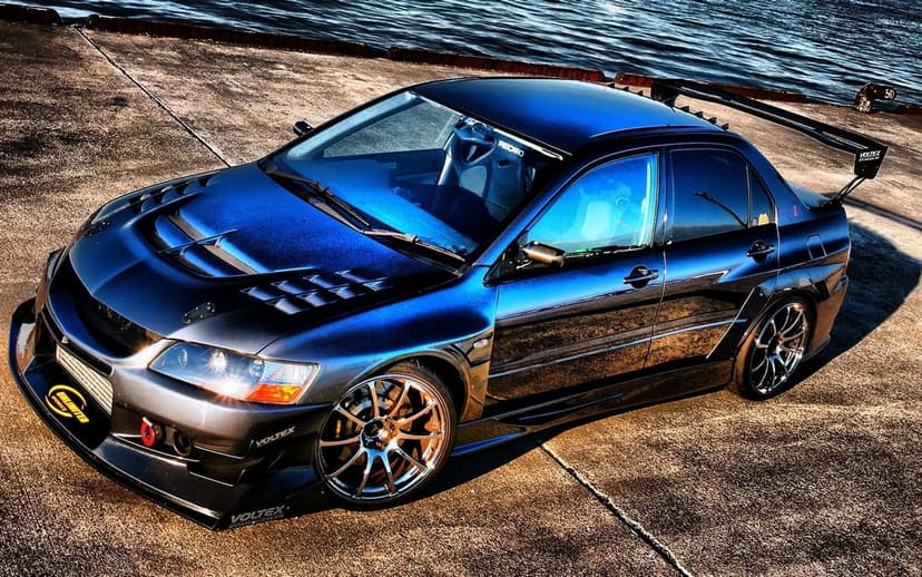 Modified blue Mitsubishi Lancer Evo IX with widebody kit and carbon hood on a pier by the sea at sunset.