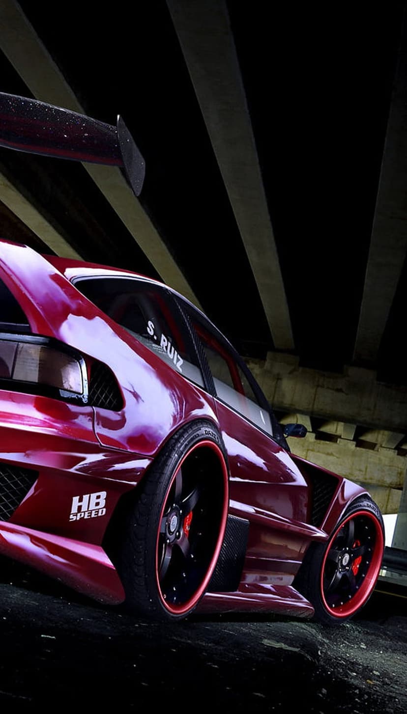 Red widebody sports car with a large spoiler and black rims parked under a concrete bridge with moody lighting.