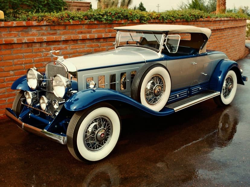 1930s blue and silver convertible classic car with white-wall tires parked on wet asphalt by a brick wall.