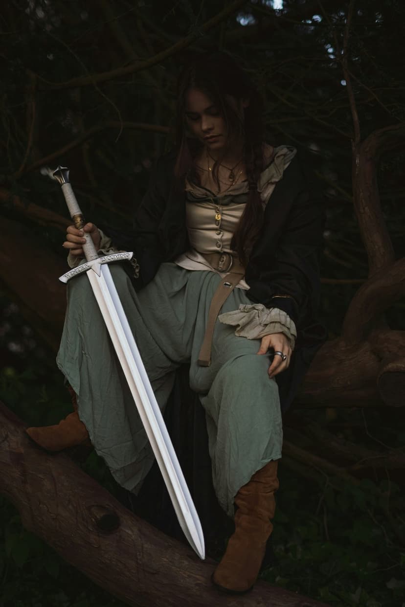 Woman in historical attire sits on a mossy branch holding a long sword within a dark, cinematic forest setting.