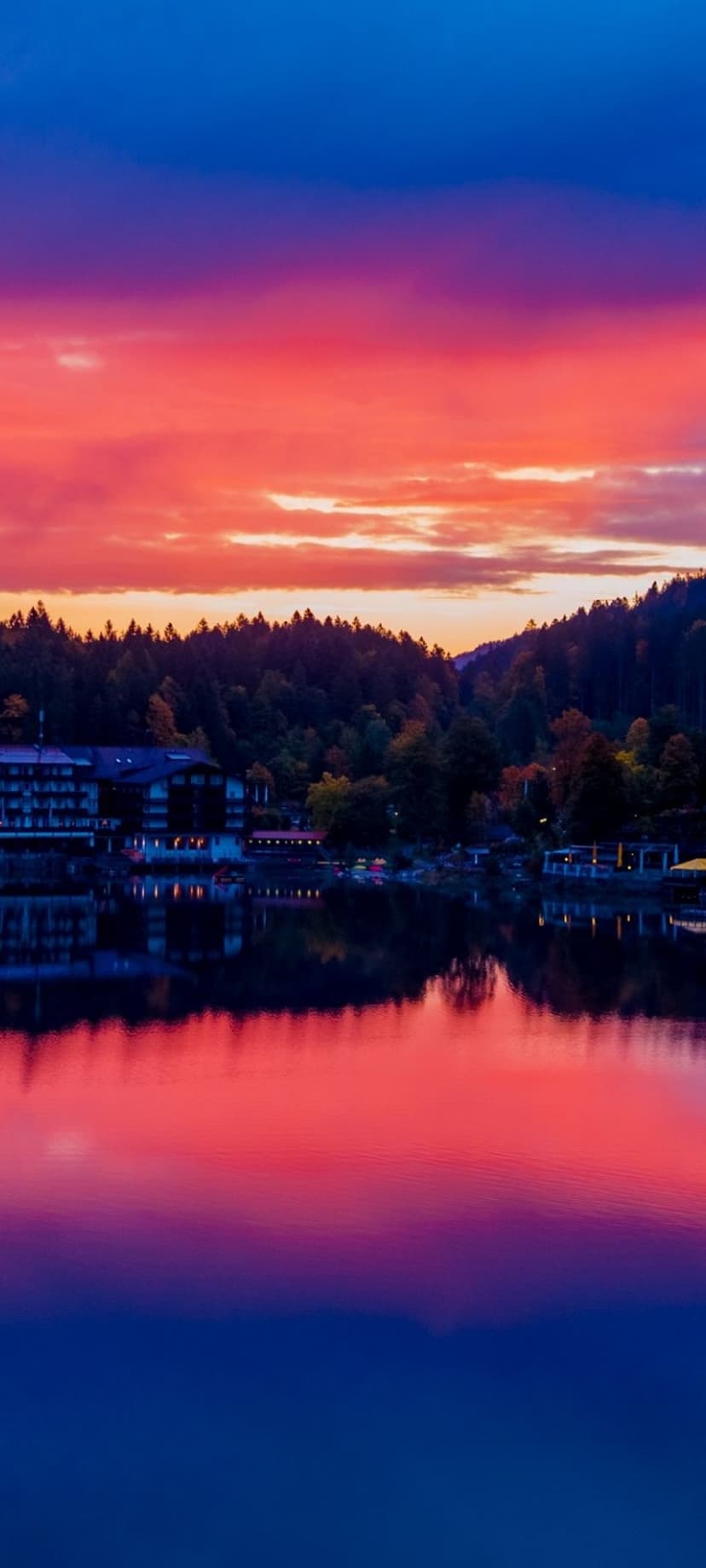 Glowing village houses reflect in a calm lake under a vibrant pink and orange sunset sky at dusk near a forest.