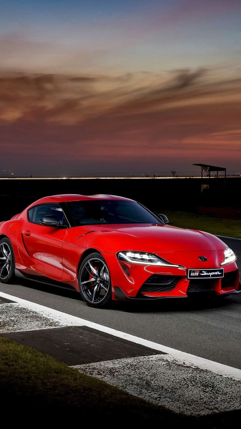 Red Toyota GR Supra sports car on a dark racetrack under a sunset sky with orange clouds and glowing headlights.