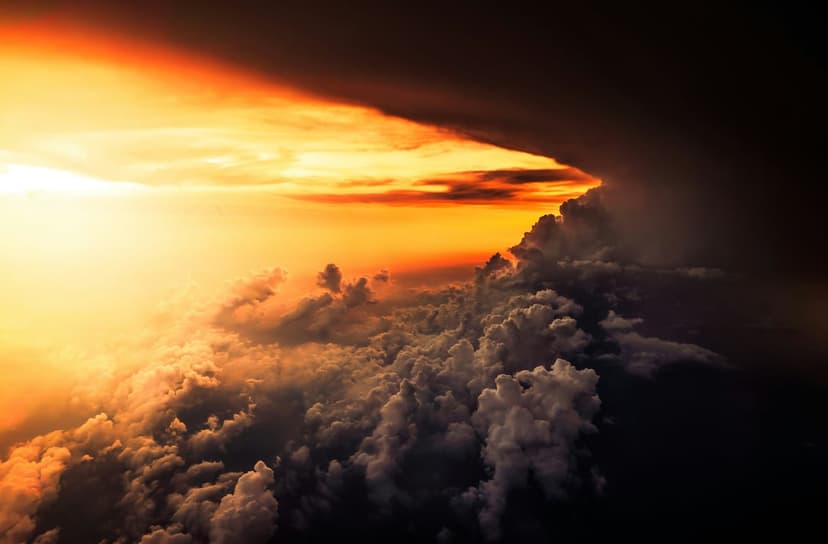 Intense orange sunlight illuminates the tops of dark, swirling storm clouds viewed from a high aerial perspective.