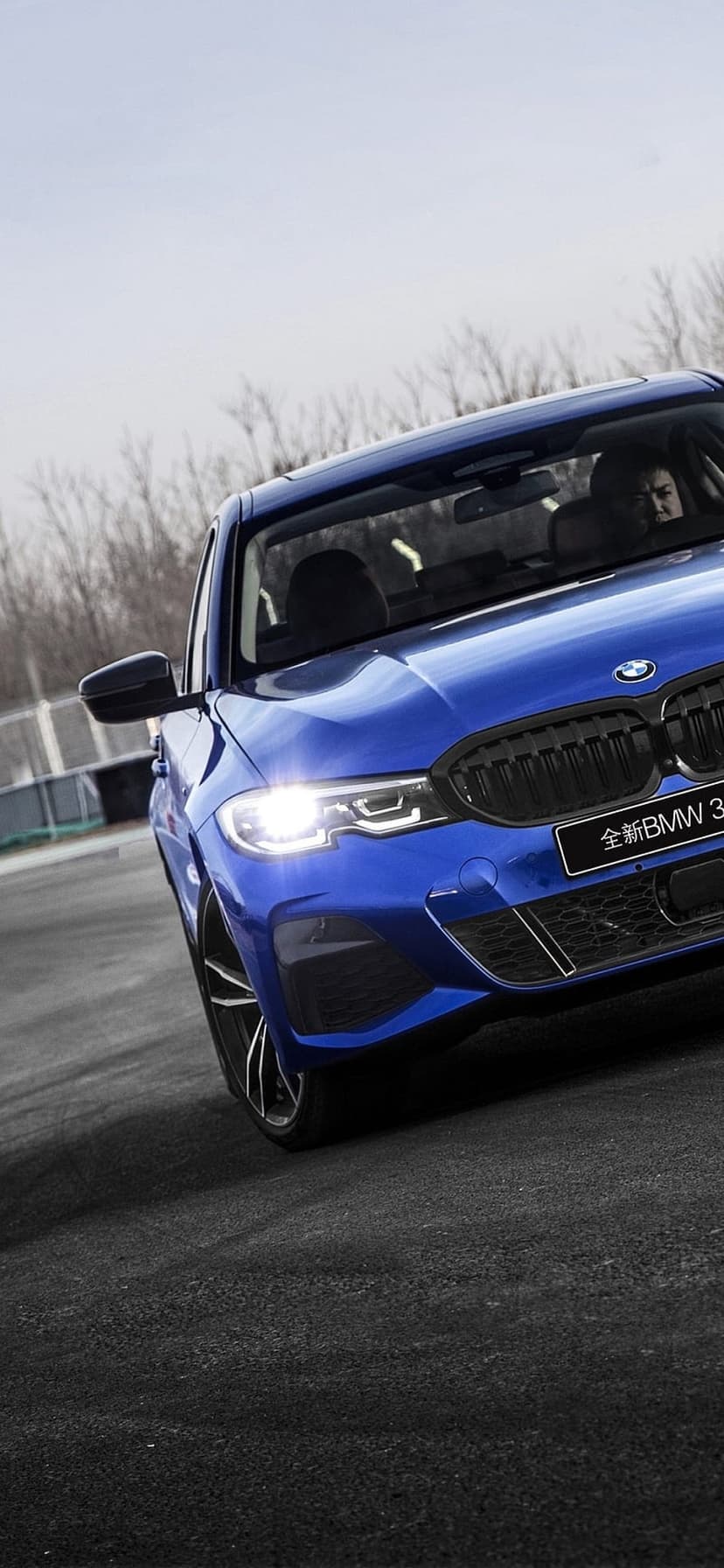 Blue BMW 3 Series sedan drives on a dark road with glowing headlights under a cloudy sky in a low-angle view.