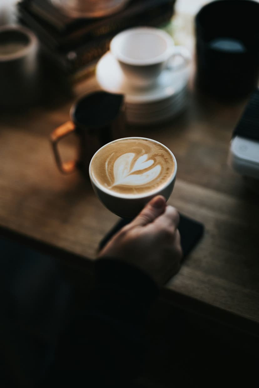 Hand holding a white cup of latte with rosette foam art over a rustic wooden table in a sunlit cafe setting.