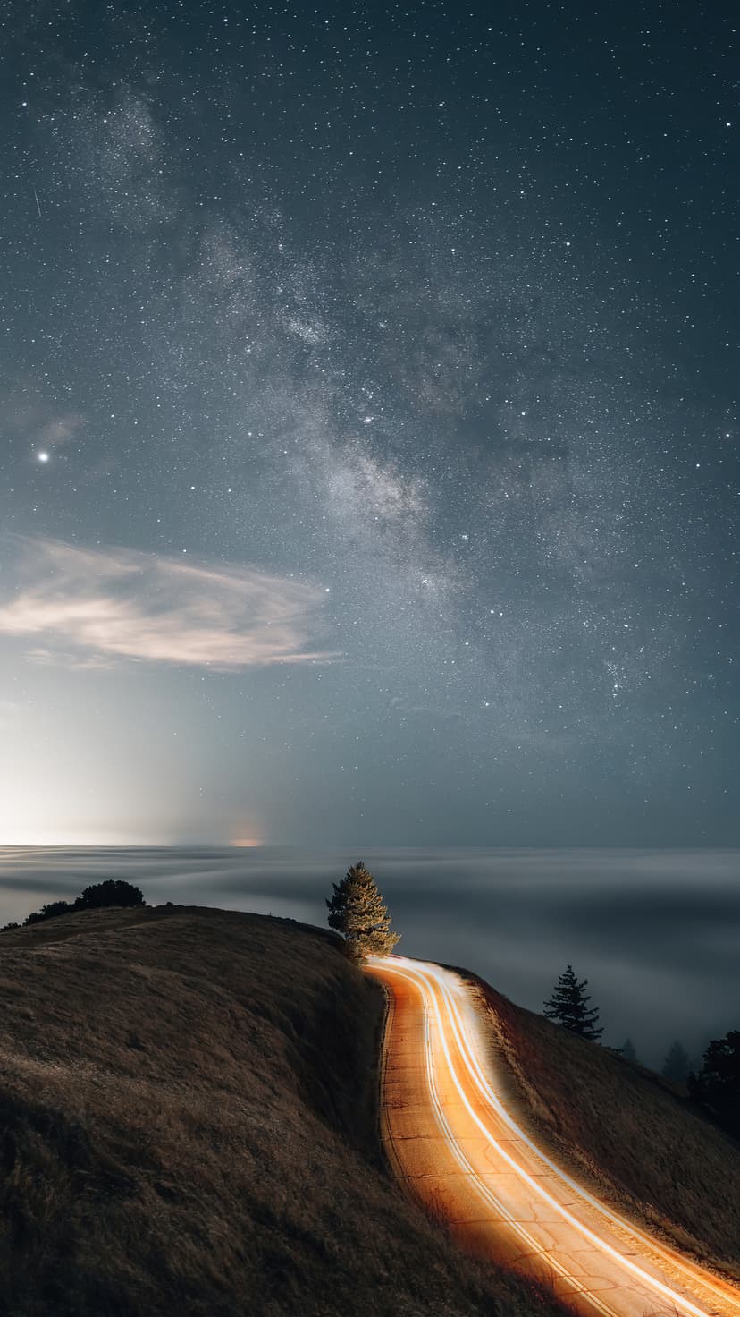 Winding road with glowing light trails under a starry Milky Way galaxy and a lone tree on a foggy hill.