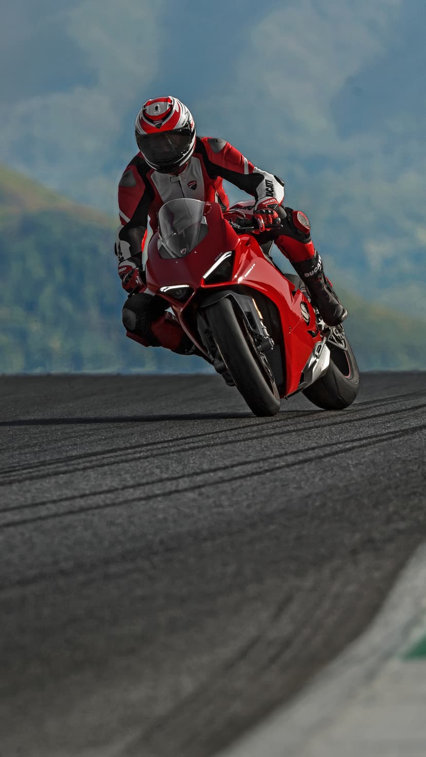 Red Ducati motorcycle rider leaning deeply into a racetrack turn with mountains and a cloudy sky in the background.