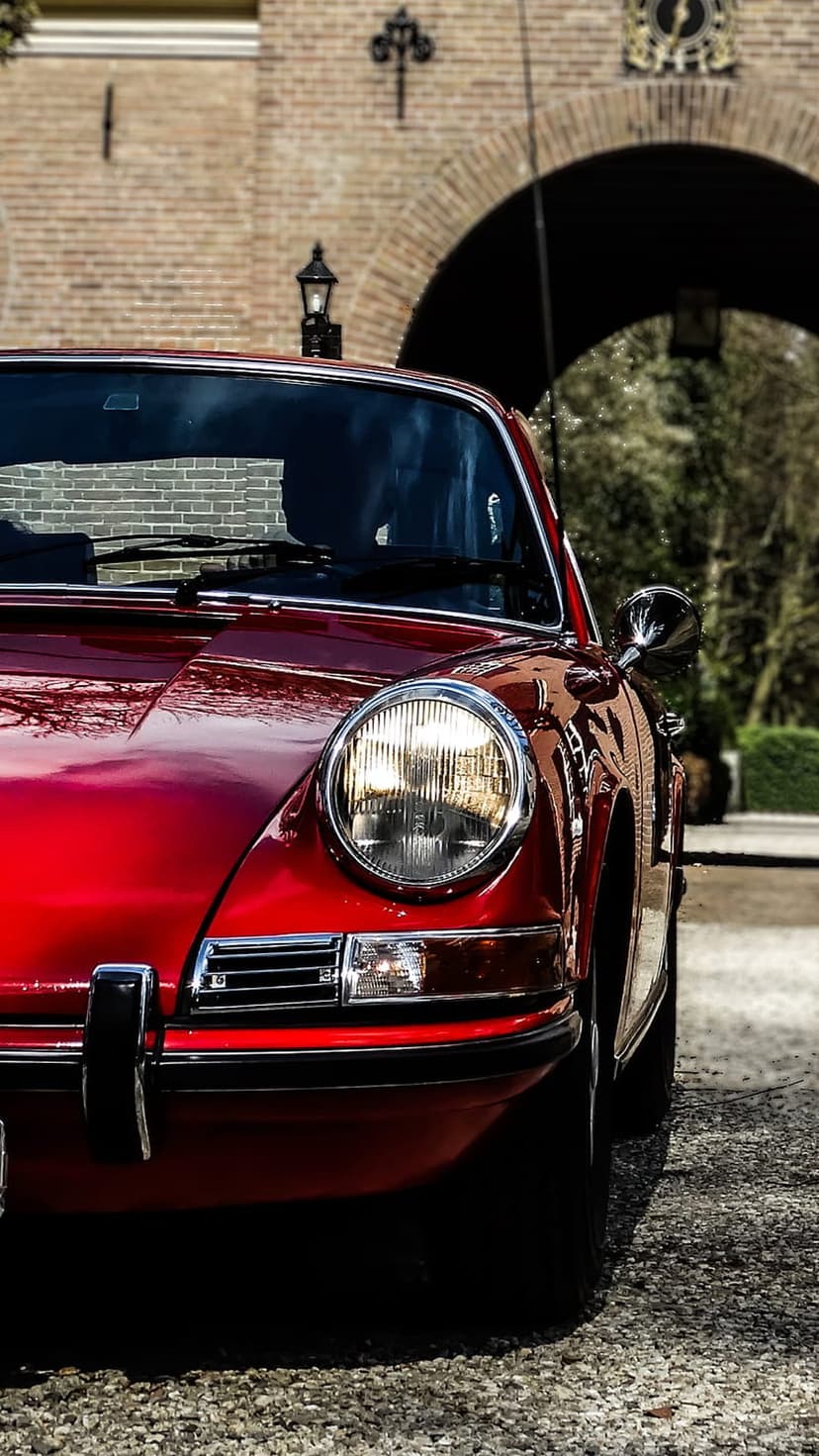 Red Porsche 911 front headlight and polished hood set against a blurred historic brick archway backdrop.