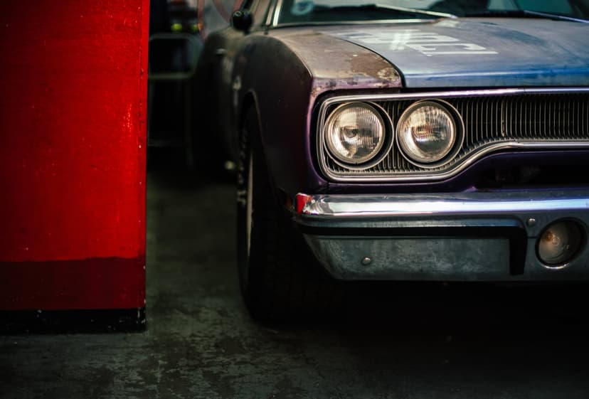 Purple muscle car headlights and rusted hood next to a red pillar in a dimly lit, atmospheric setting.