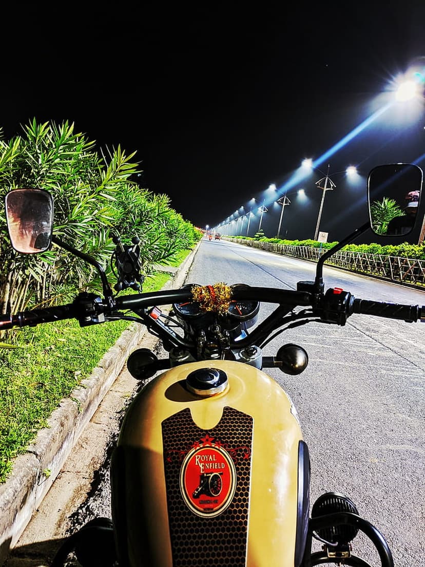 Royal Enfield motorcycle handlebars and fuel tank illuminated by warm streetlights on a dark asphalt road at night.